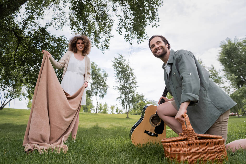 low angle view of cheerful woman in summer clothes holding blanket near boyfriend with wicker basket in park - Foto, Imagem