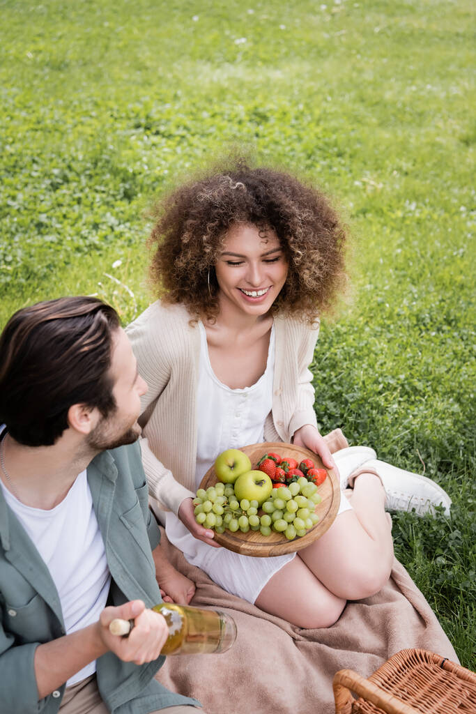 Kıvırcık kız arkadaşının yanında elinde şarap şişesi olan mutlu bir adam tahta ve meyvelerle oturuyor. - Fotoğraf, Görsel