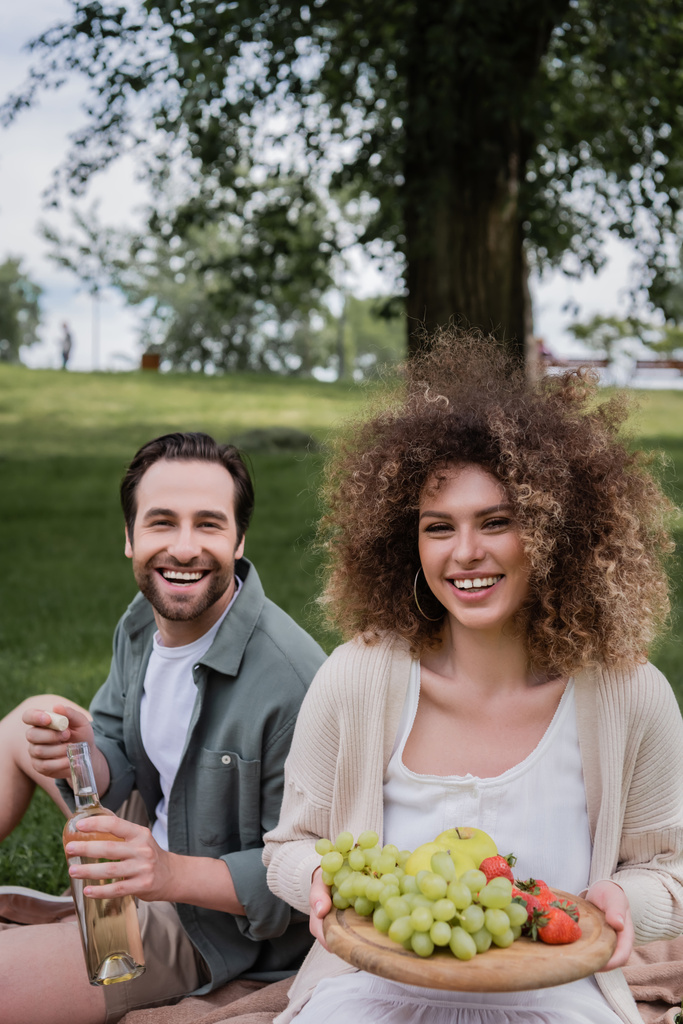 happy man opening bottle of wine near smiling curly girlfriend sitting with fruits during picnic - Foto, immagini