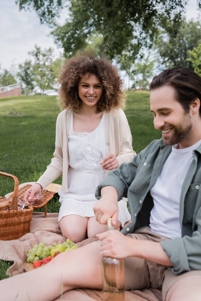 happy curly woman looking at man opening bottle of wine during picnic - Photo, image