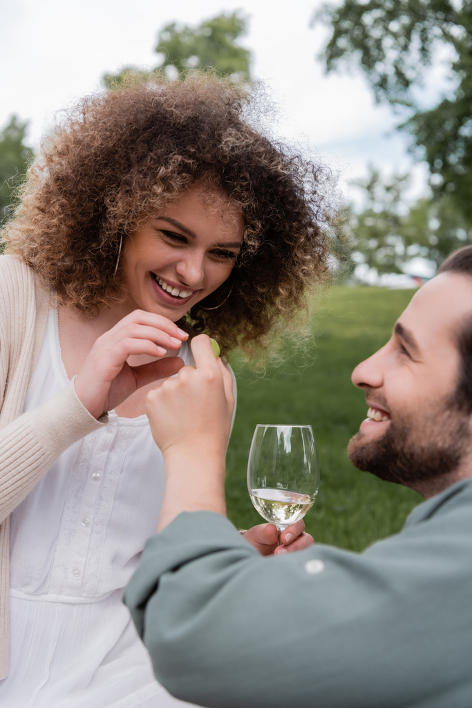 happy man with glass of wine holding fresh grape near cheerful girlfriend during picnic - Zdjęcie, obraz