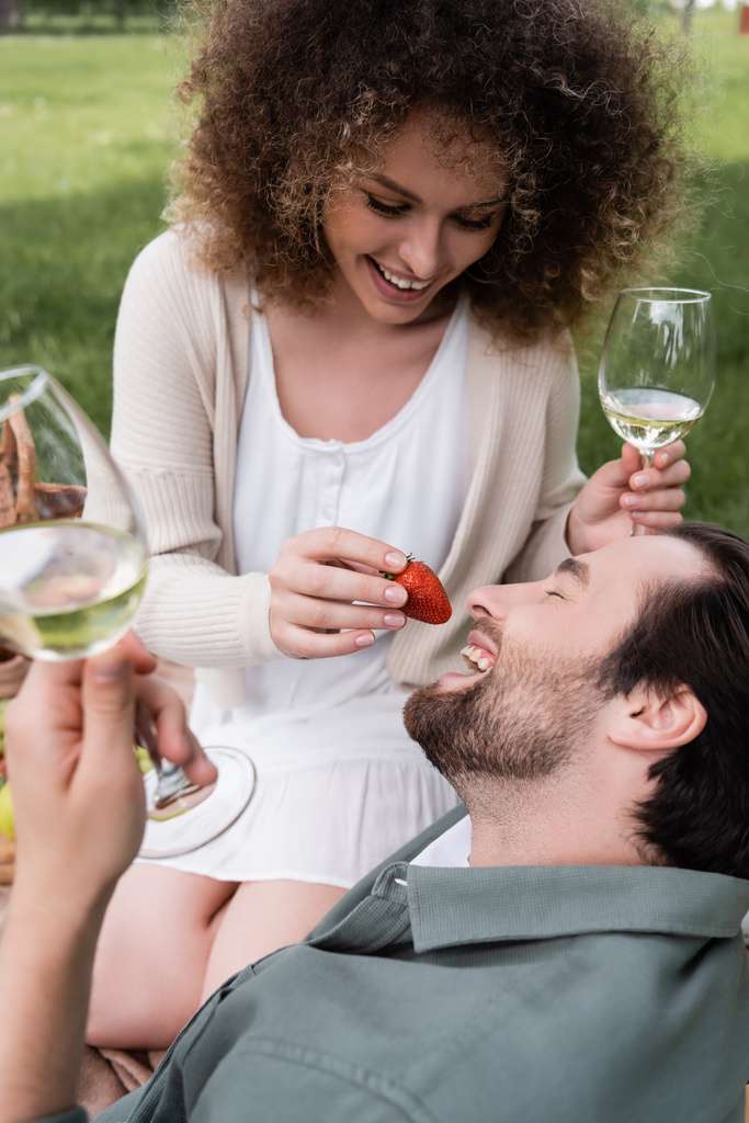 happy curly woman holding glass of wine and feeding boyfriend with fresh strawberry during picnic - Foto, afbeelding