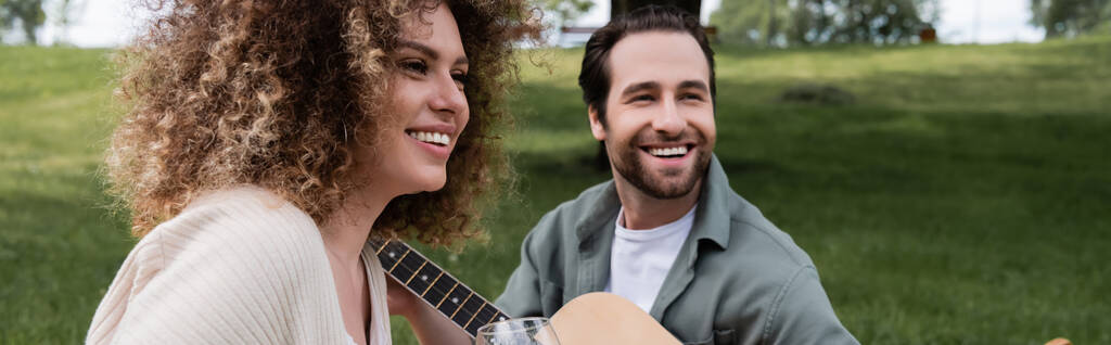 romantic man playing acoustic guitar near curly woman during picnic, banner - 写真・画像