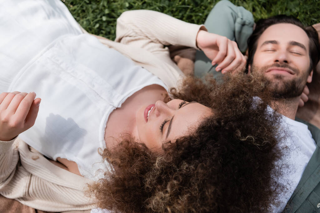 vista de ángulo alto de la mujer rizada acostada en novio complacido durante el picnic en el parque de verano - Foto, imagen