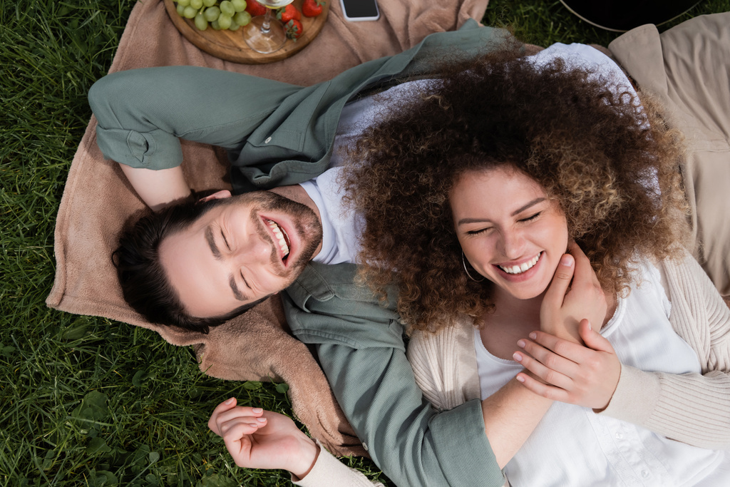 top view of curly woman lying on joyful boyfriend during picnic in summer park - Fotó, kép