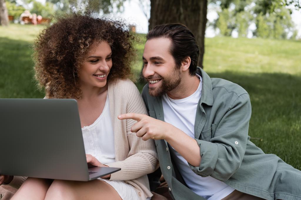 счастливый мужчина показывает пальцем на ноутбук рядом с веселой женщиной  - Фото, изображение