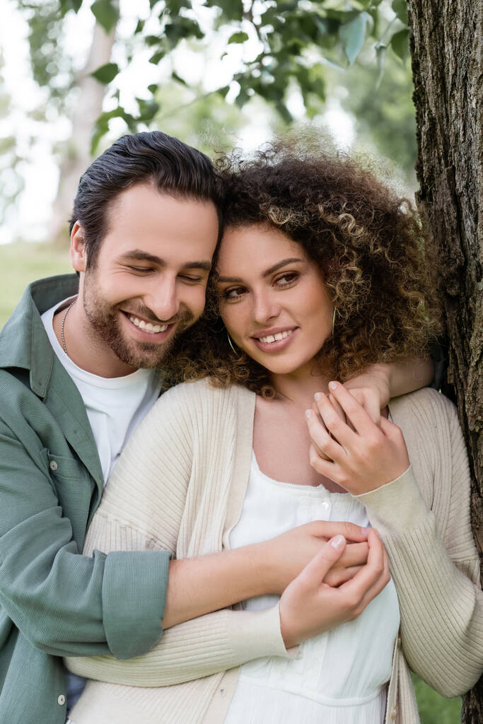 happy man embracing curly and positive woman near tree trunk  - Foto, imagen