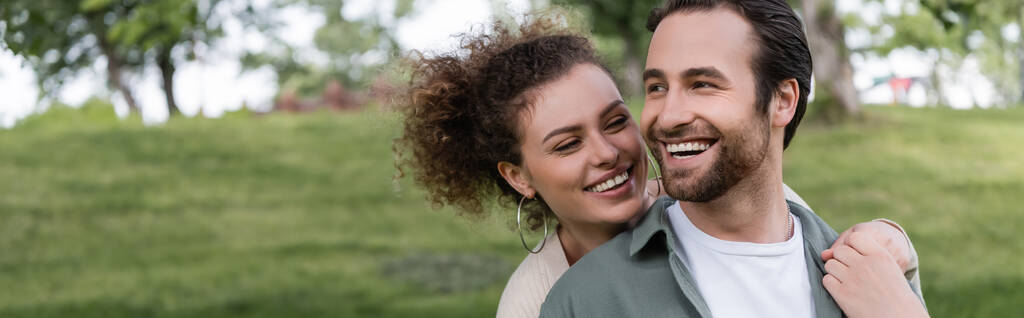 довольная и кудрявая женщина обнимает парня сзади в зеленом летнем парке, баннер - Фото, изображение