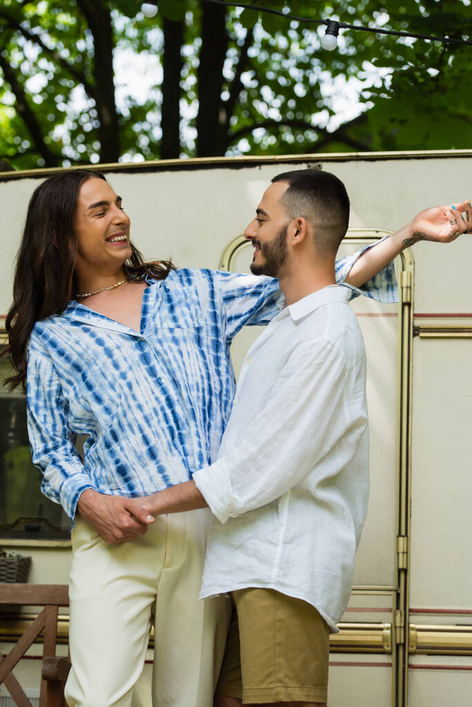 cheerful gay couple smiling while hugging during camping near van  - Foto, Bild