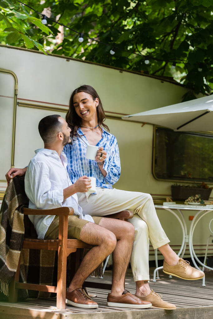 felice gay uomo holding tazza di caffè mentre seduta con fidanzato vicino van durante viaggio in estate - Foto, immagini