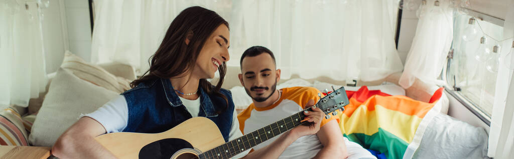 happy and tattooed gay man playing acoustic guitar near boyfriend on bed in modern van, banner - Photo, Image