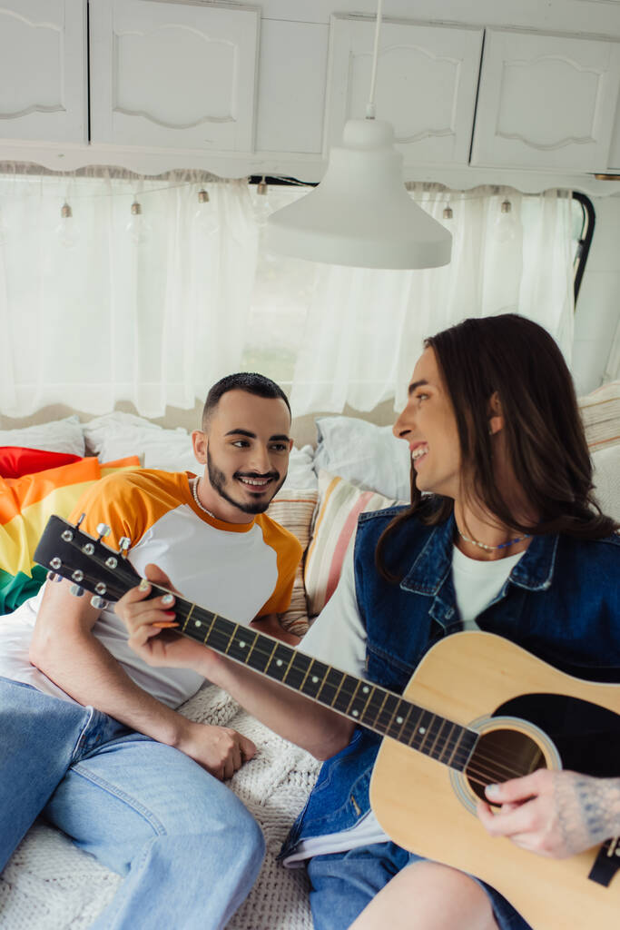 happy and tattooed gay man playing acoustic guitar near boyfriend and lgbt flag on bed in van  - Valokuva, kuva