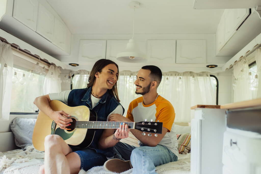 happy gay man playing acoustic guitar near bearded boyfriend on bed in modern van  - Foto, afbeelding