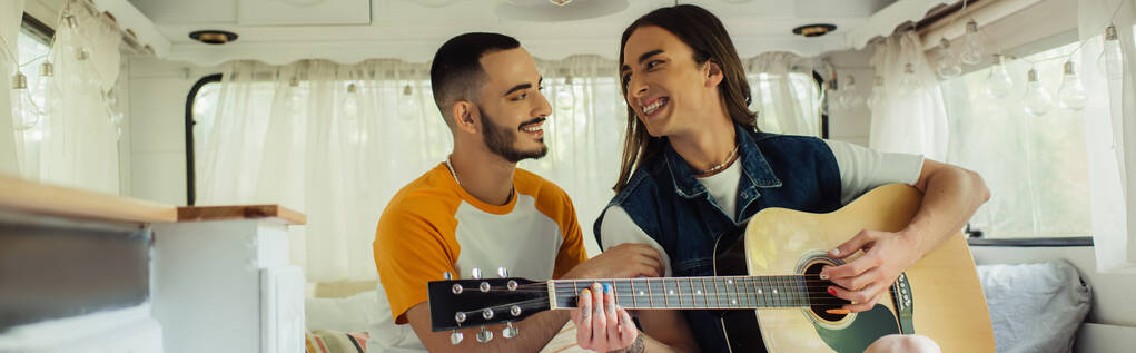 glücklich tätowierter schwuler Mann spielt Akustikgitarre neben Freund in modernem Van, Banner - Foto, Bild