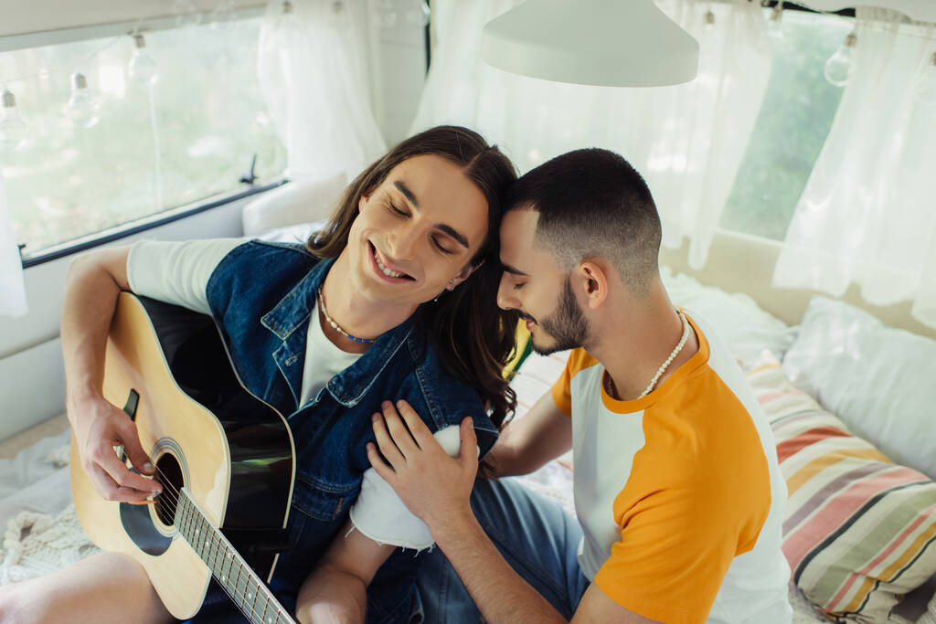 high angle view of happy gay man with long hair playing acoustic guitar near boyfriend on bed in modern van  - Fotografie, Obrázek