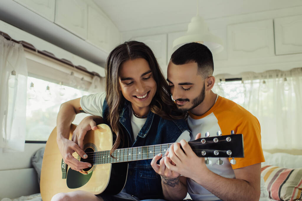 Улыбающийся гей с длинными волосами играет на акустической гитаре рядом с счастливым парнем в современном фургоне  - Фото, изображение
