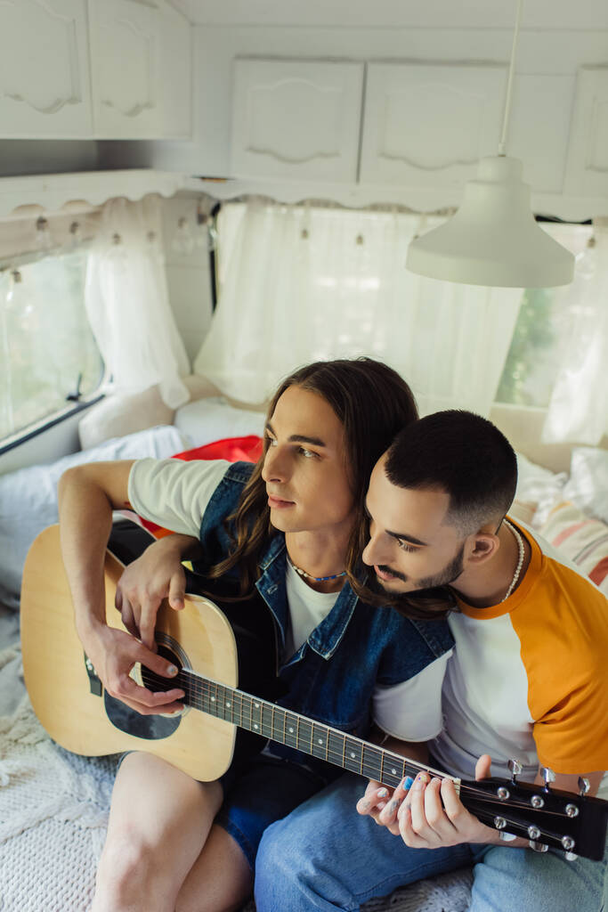 high angle view of gay man with long hair playing acoustic guitar near boyfriend on bed in modern van  - Foto, Imagen
