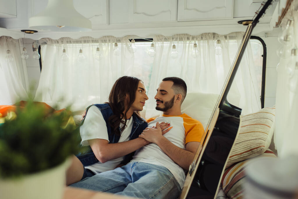gay man with long hair hugging bearded boyfriend on bed near guitar in modern van  - Fotó, kép