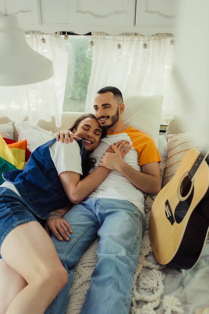 smiling gay man with tattoo lying in bed with bearded boyfriend near lgbt flag in modern van  - Foto, Imagen