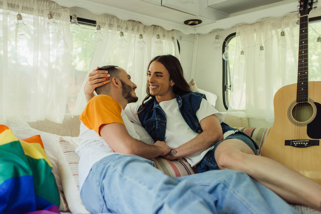 smiling gay man with tattoo looking at bearded boyfriend on bed near lgbt flag and acoustic guitar in modern van  - Foto, immagini