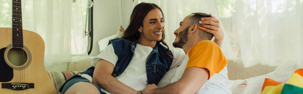 smiling gay man with tattoo looking at bearded boyfriend on bed near lgbt flag and acoustic guitar in van, banner - Zdjęcie, obraz