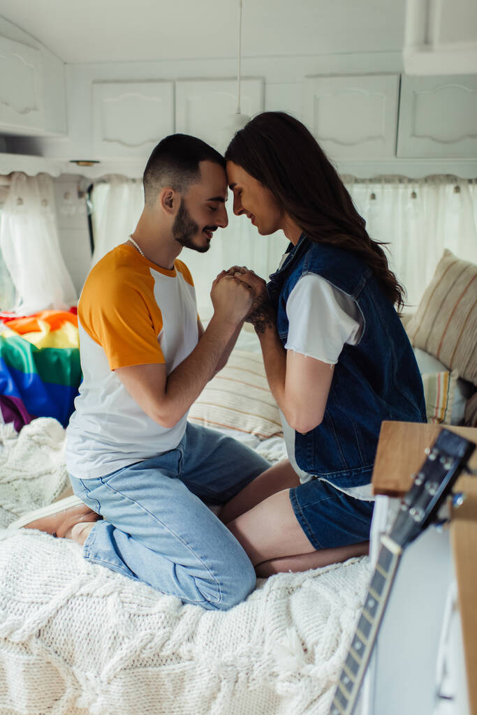 vista lateral de homem gay com tatuagem de mãos dadas com namorado barbudo na cama em van  - Foto, Imagem