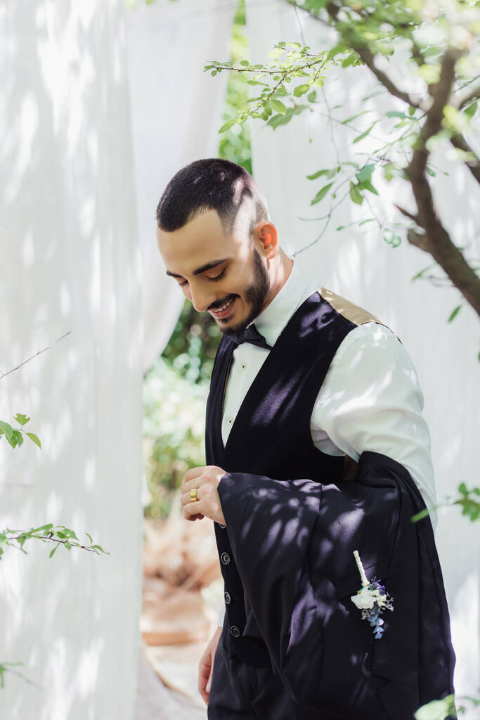 novio barbudo feliz en ropa formal mirando el anillo de bodas de oro en el dedo - Foto, imagen