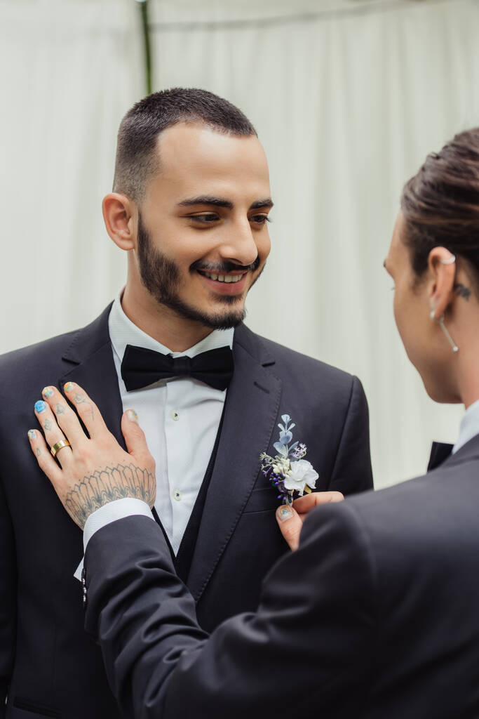 татуйований гей чоловік регулює бутоньєрку на костюмі щасливого бородатого нареченого
 - Фото, зображення