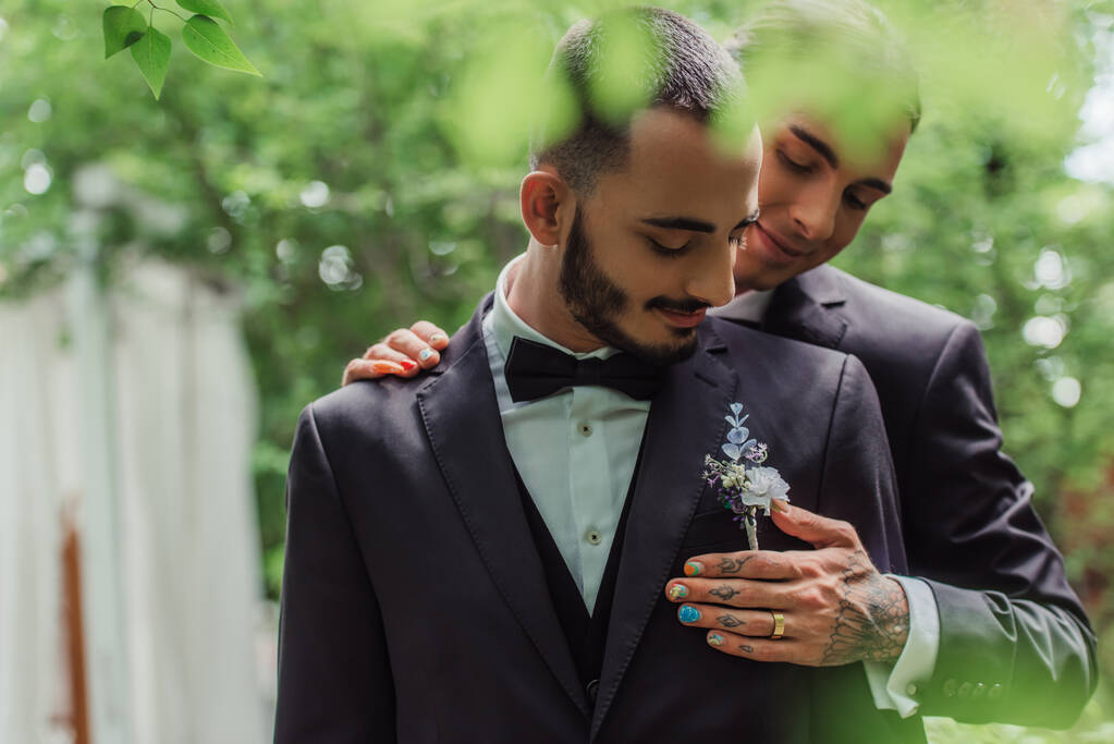 cheerful gay man with wedding ring hugging groom in formal wear with boutonniere - Foto, Bild