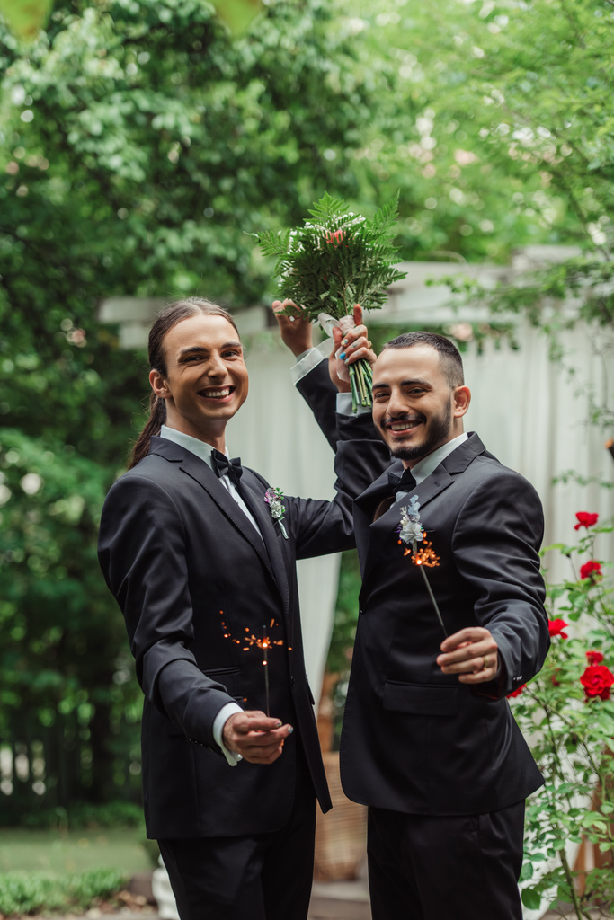 glücklicher schwuler Mann in formeller Kleidung mit Brautstrauß und Wunderkerze in der Nähe des Bräutigams im grünen Park - Foto, Bild