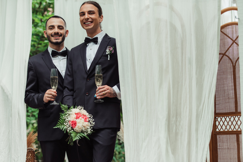 cheerful gay newlyweds in suits holding glasses of champagne on wedding day  - Fotó, kép