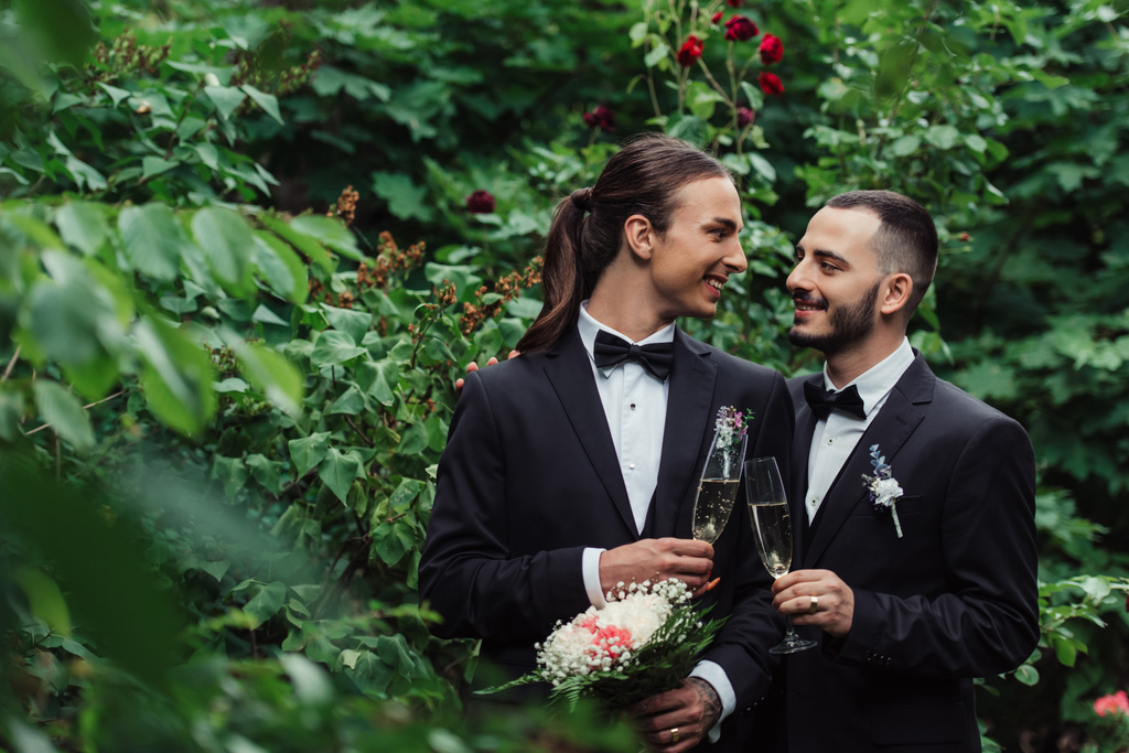 happy gay couple in suits holding glasses of champagne in green park - 写真・画像