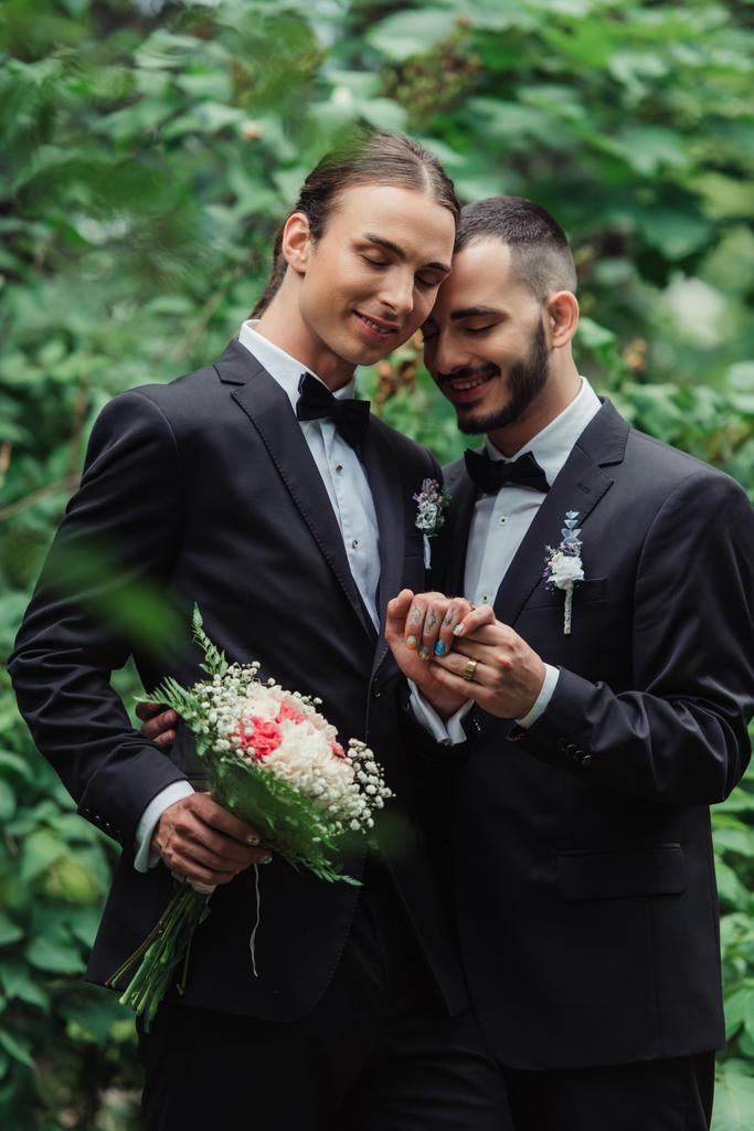 joyful gay couple in suits holding hands near wedding bouquet in green park - Photo, Image