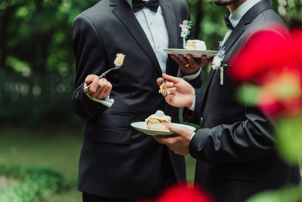 vista ritagliata di sposi gay in abbigliamento formale tenendo torta nuziale sul piatto  - Foto, immagini