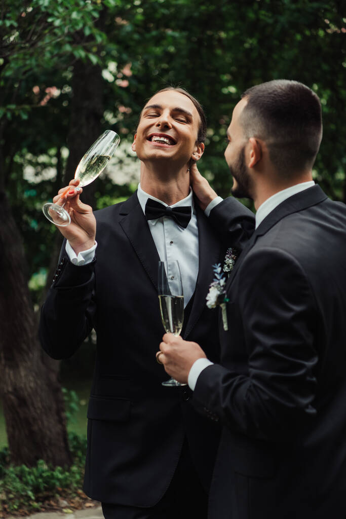 позитивные новобрачные геи в костюмах, держащие бокалы с шампанским в день свадьбы  - Фото, изображение