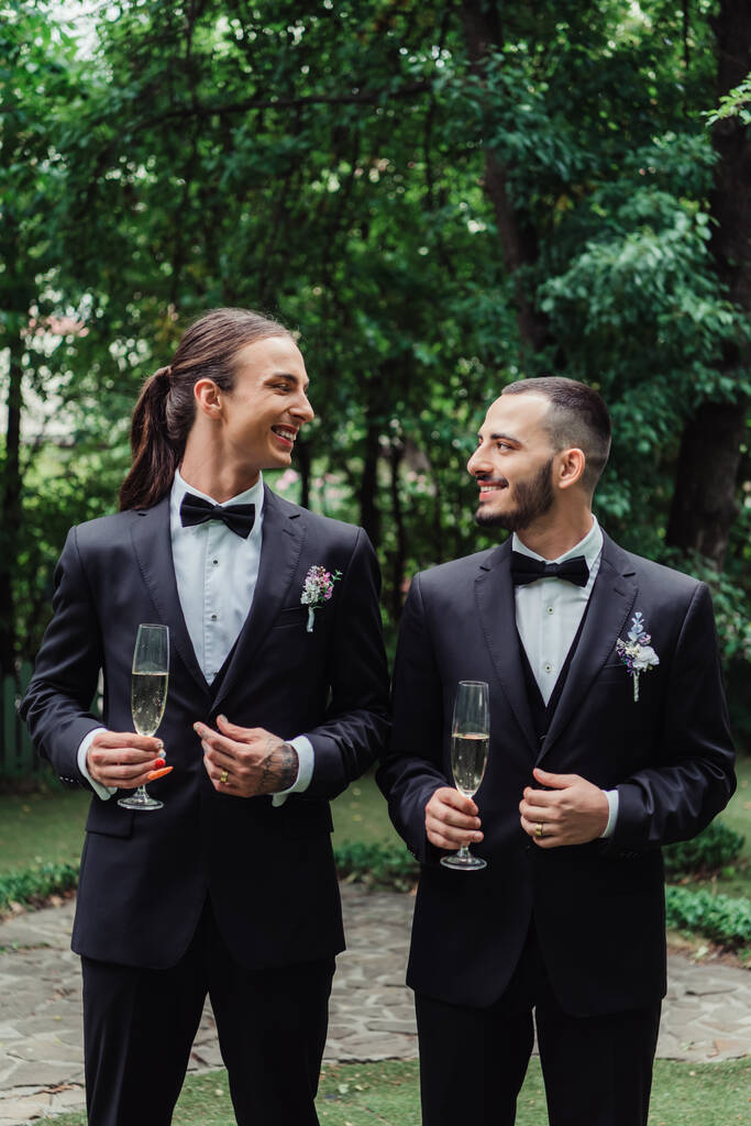 pleased gay newlyweds in suits holding glasses with champagne on wedding day  - Foto, afbeelding