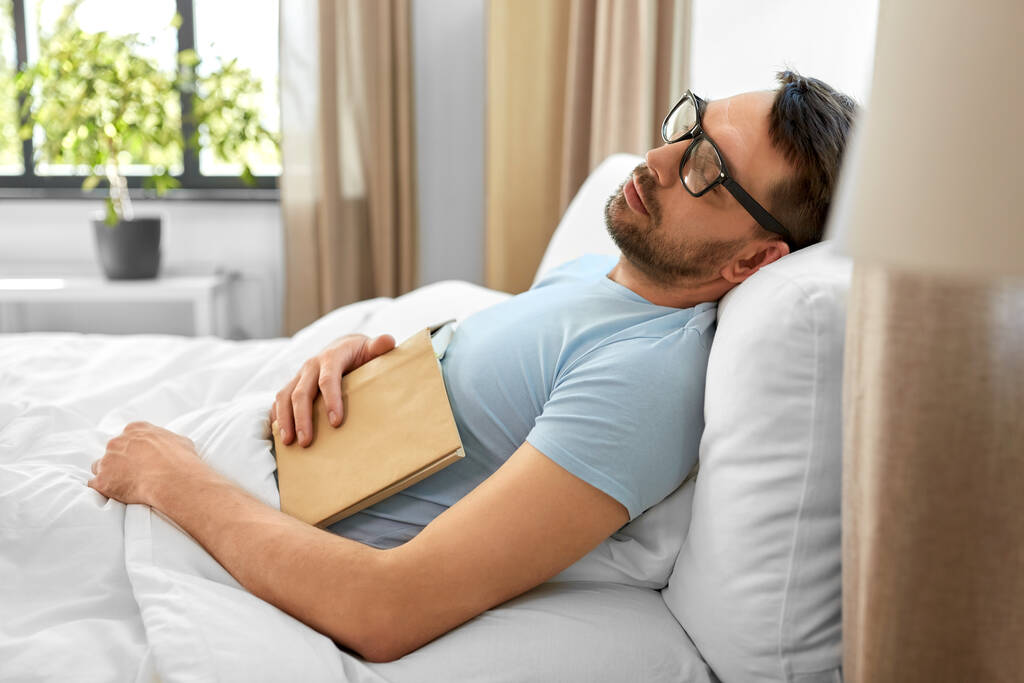 ludzie, koncepcja snu i odpoczynku - mężczyzna w okularach z książką śpiący rano w łóżku w domu - Zdjęcie, obraz