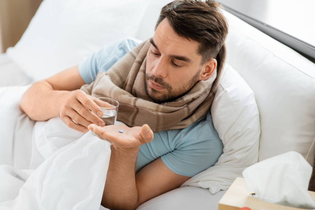 ludzie i problem zdrowotny koncepcja - nieszczęśliwy chory człowiek z pigułki lekarskiej i szklanki wody leżącej w łóżku w domu - Zdjęcie, obraz