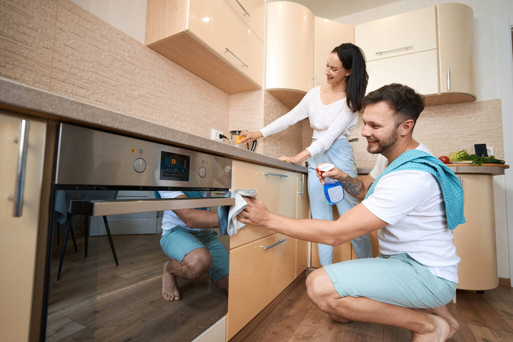 Χαμογελώντας αρσενικό σκούπισμα συρτάρια ντουλαπιών κουζίνας με πανί και υγρό καθαρισμού ψεκασμού, ενώ η γυναίκα χρησιμοποιεί σφουγγάρι στο βραστήρα - Φωτογραφία, εικόνα