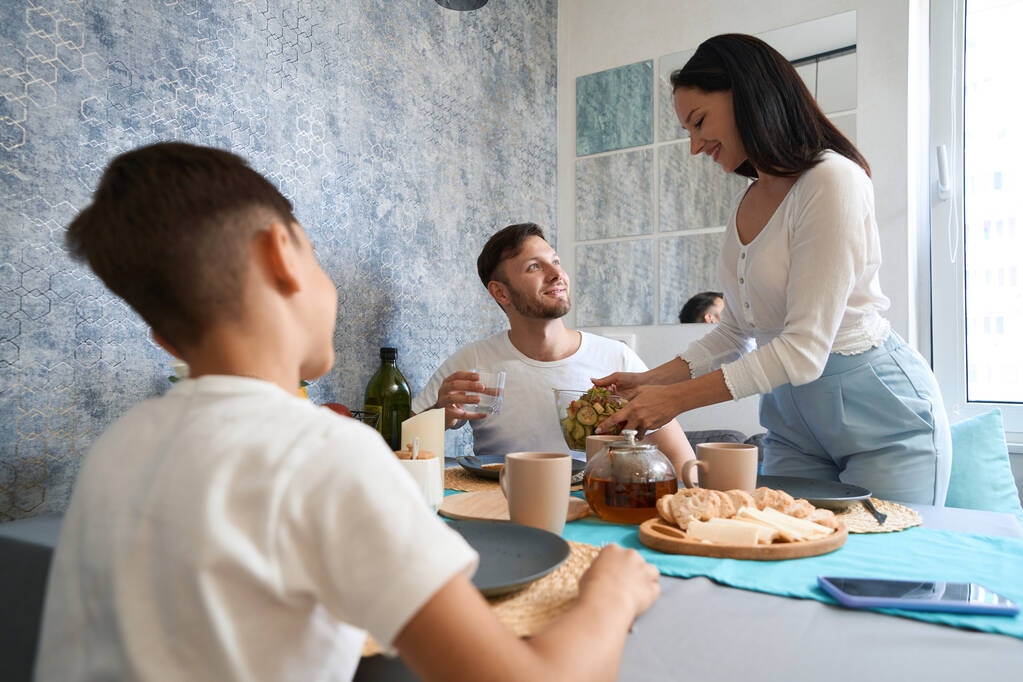 Nainen hymyilee laittaessaan kasvissalaattia miehensä ja poikansa lautasille perheen aterian aikana. - Valokuva, kuva
