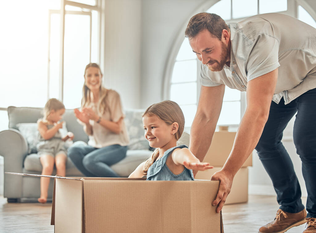A happy mature caucasian father pushing his daughter in a box while her mother and sister sit on the sofa in the lounge at home. Man and girl having fun, playing games at home while enjoying family - Fotoğraf, Görsel