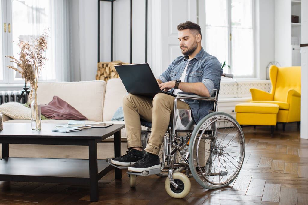 Baard blanke man met behulp van rolstoel typen op draadloze laptop in de woonkamer. Jonge mannelijke freelancer met speciale behoeften werken op afstand van huis. - Foto, afbeelding