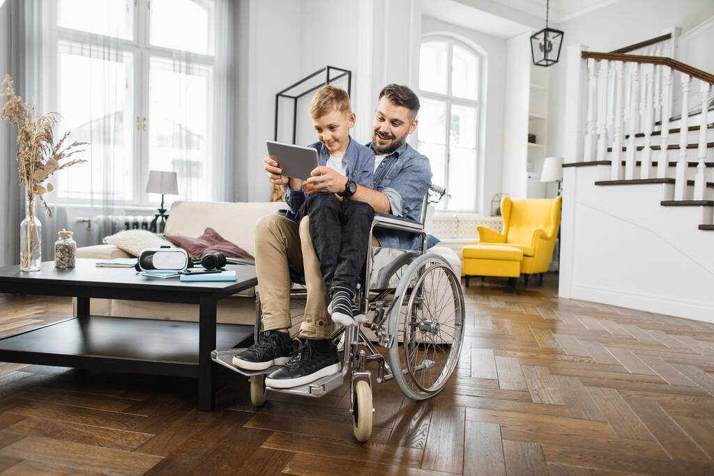Usmívající se táta se speciálními potřebami sedí na invalidním vozíku a drží syna na kolenou, zatímco hraje na digitálním tabletu. Pozitivní rodina používající přenosné zařízení pro hraní dětských vzdělávacích her. - Fotografie, Obrázek