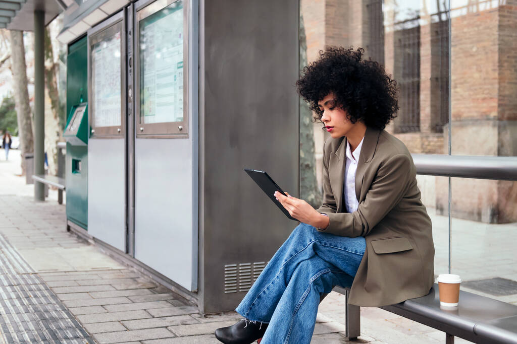 young latin business woman sitting at the bus stop using her tablet, concept of technology and urban lifestyle - Zdjęcie, obraz