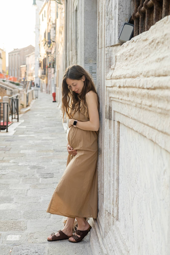 Uśmiechnięta ciężarna kobieta dotykająca brzucha w pobliżu budynku na ulicy w Wenecji  - Zdjęcie, obraz
