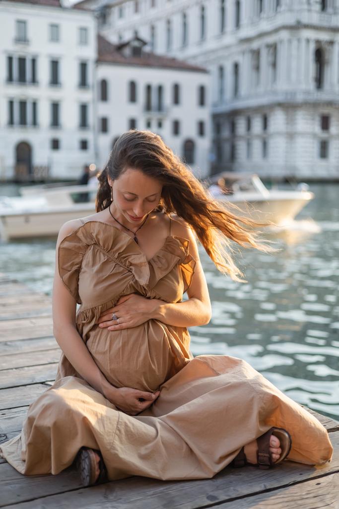 Smiling pregnant woman sitting on wooden pier in blurred Venice  - Foto, Imagem