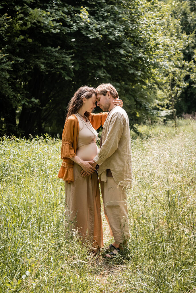 Vista lateral de la mujer embarazada abrazando marido en el campo  - Foto, imagen