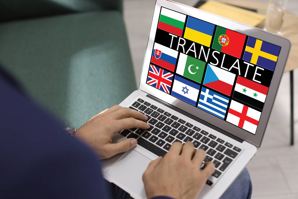 Translator using modern laptop with images of different flags on screen in office, closeup - Foto, Bild