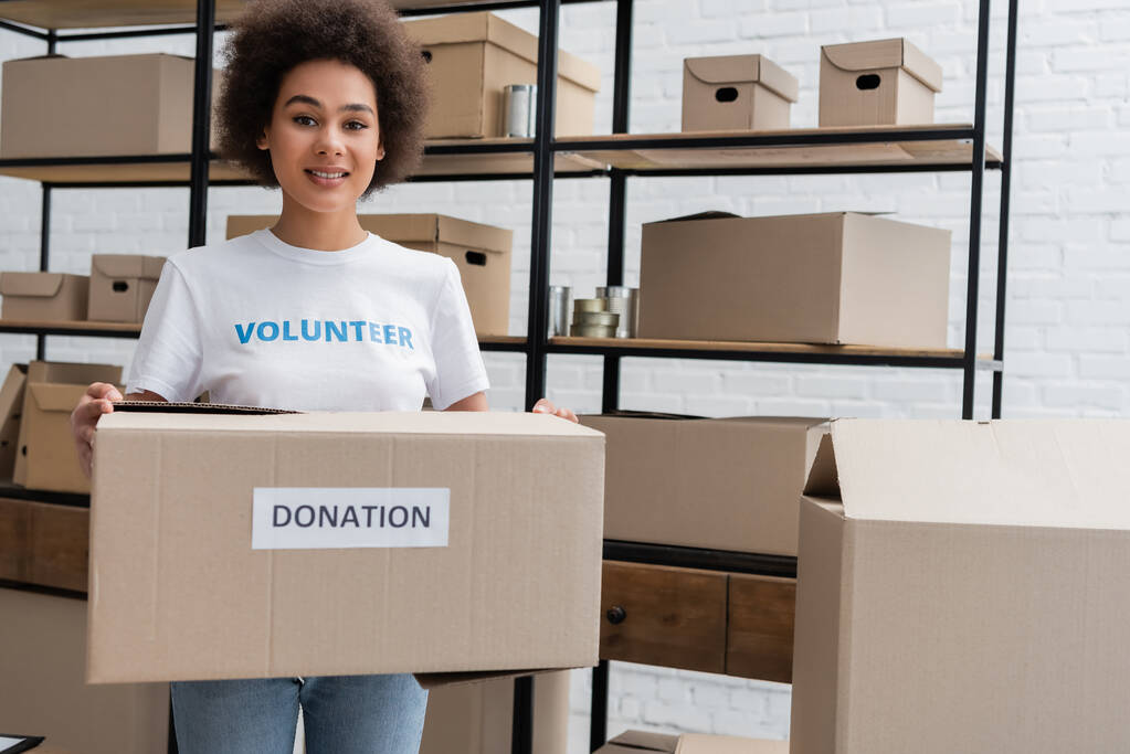 african american volunteer smiling at camera while holding donation box in charity warehouse - Foto, Bild