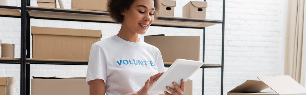 happy african american volunteer using digital tablet while working in donation center, banner - Foto, Bild